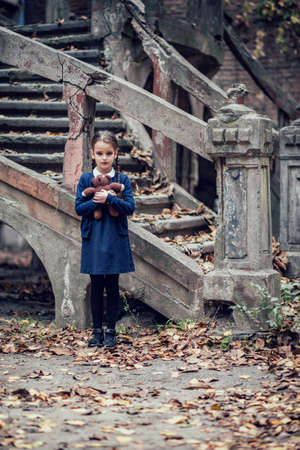 Beautiful little brunette girl with with pigtails, dressed in dark blue standing near mystic abandoned building with gothic stairs and holding handmade bear toy. Halloween horror, ghost or spirit of child in twilightの写真素材
