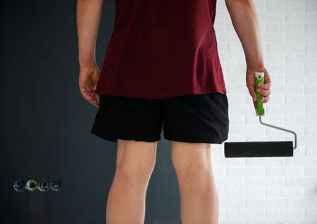 Roller Wall Painting. Man standing near black wall and holding roller, back view. Worker ready to paint, home renivation. Painter at work. Home renovationの写真素材