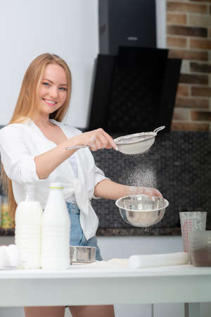 Young woman standing in the kitchen at home and cooking with enjoyment bakery products of flour, milk, cocoa, sugar and fruits. Funny portrait of happy girl chef sifts flourの写真素材