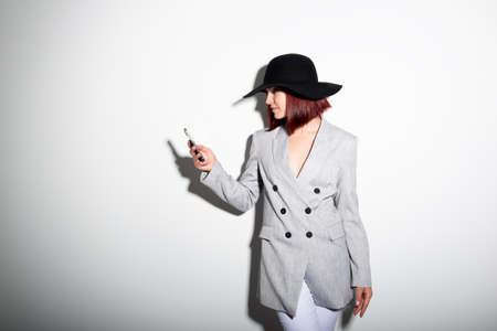 Young stylish woman using smartphone, indoor portrait over white. Fashion modern girl wearing grey jaket and black hat texting on her mobile phoneの写真素材
