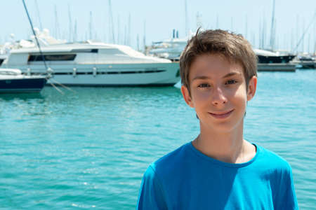 Portrait of teen boy over aquamarine water with sailing ships in port of Alicante, Spain.