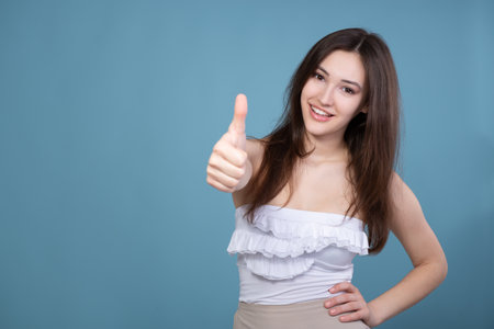 Attractive smiling young woman with thumbs up gesture demonstrating success and victory in business and life, isolated on blue backgroundの写真素材