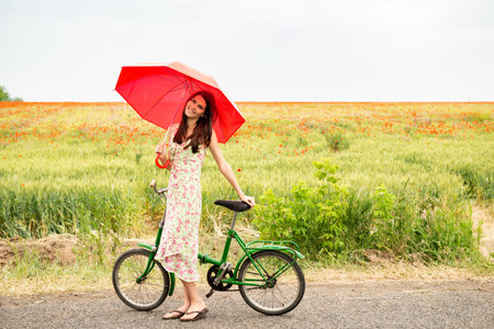 Young beautiful woman with bicycle walks on a poppy field, summer outdoor. Attractive girl holding red umbrella with retro bikeの写真素材