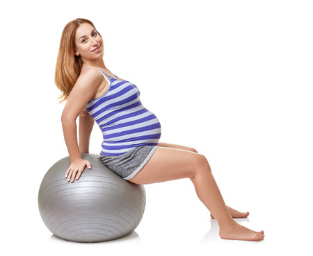 Pregnant woman with big belly doing sport exercises on fit ball at studio over white background. Pregnancy and healthy active lifestyleの写真素材