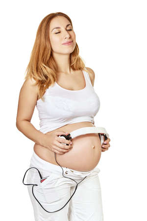 Pregnant woman with with headphones on her big belly posing at studio over white background. Pregnancy.の写真素材