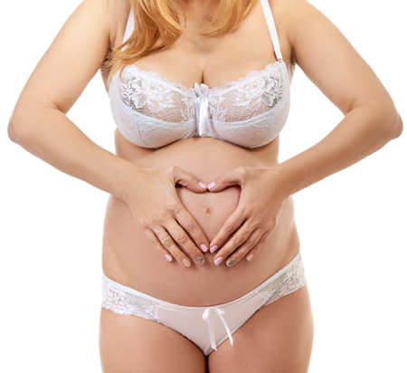 Pregnant woman touching her big belly with hands like heart sign, symbol maternity and mother care, studio shot over white background. Pregnancy.の写真素材