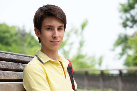Young handsome 15 years old teen boy wearing yellow t-shirt sitting on a bench in the park, summer outdoorの写真素材