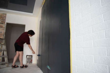 Roller Wall Painting. Man standing near black wall and holding roller, back view. Worker paints wall, home renivation. Painter at work. Home renovationの写真素材