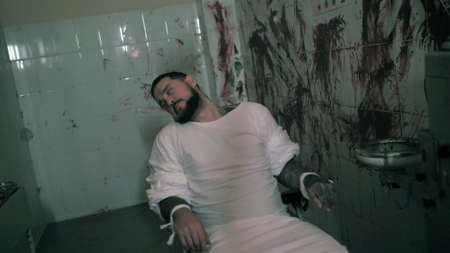 Crazy man in straitjacket tied to a chair, killer psychopath in a room with bloodied walls overdose