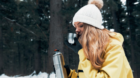 A girl in a yellow jacket pours tea from a vacuum flask in the winter in the forestの写真素材