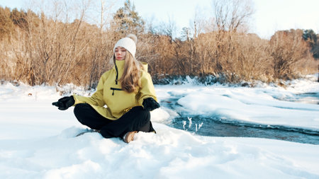 The girl sits in the lotus position in the winter in the forest and looks at the river. Front viewの写真素材