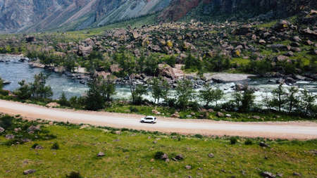A white SUV car is driving along an empty country road in the huge Katu-Yaryk canyon Chulyshman Valley with a large mountain bubbling river. Altai, Siberia, Russiaの写真素材