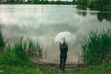 Girl with an umbrella from the back. Umbrella transparent, rainy weather. Beautiful nature, a lake and many trees.の写真素材