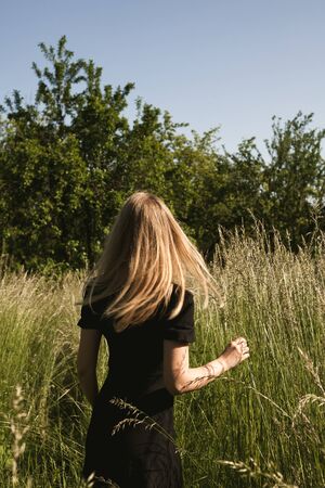 Girl in a black dress is walking in nature. Blonde in motion in the field. A young girl enjoy the...