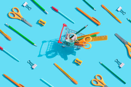 Back to school. Miniature supermarket trolley with stationery inside: scissors, pens, pencils, paper clips, ruler, tape. The same objects are spread around. Blue background, top view, flat lay.の写真素材