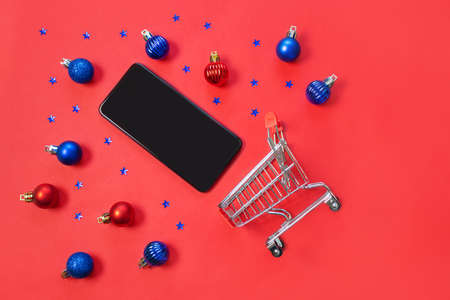 Supermarket trolley, smartphone and blue and red Christmas balls and stars on a red background. Minimalism, copy space, flat lay. The concept of New Year and Christmas sales, shopping, online shopping.の写真素材
