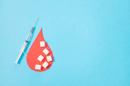 World Diabetes Day. A large paper drop of blood and a syringe on a blue background, around pieces of refined sugar. Flat lay, creative concept of health, blood sugar, minimalism.の写真素材