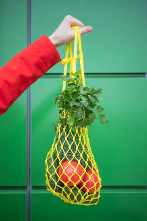 Yellow string bag with cucumbers, tomatoes, bananas and herbs in hand close-up. Bright photo in red, yellow, green tones. Sustainability, zero waste, plastic free concept, vegetarianism, healthy food.の写真素材