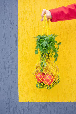 Yellow string bag with cucumbers, tomatoes, bananas and herbs in hand close-up. Bright photo in red, yellow, green tones. Sustainability, zero waste, plastic free concept, vegetarianism, healthy food.の写真素材