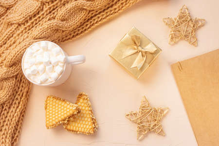 Cozy home still life: white cup with cocoa with marshmallows, cookie, chunky knit scarf, golden gift...
