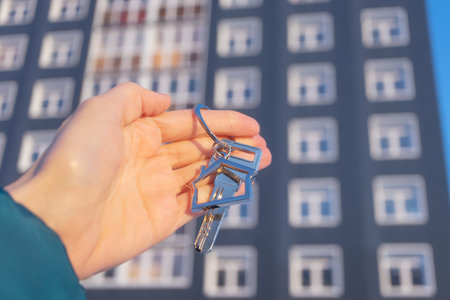 Close-up of key with keychain in shape of house in hand. On the background is multi-storey residential building. Concept of buying and insuring real estate, apartments, mortgages. Copy space.の写真素材