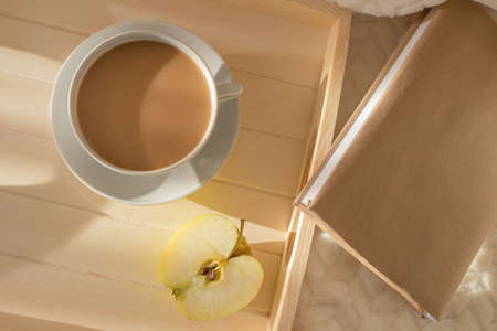 Wooden tray with a cup of coffee and an apple on a light blanket. There is a book nearby. View from above. The concept of a morning, weekend, relaxing home. Bright sunshine.の写真素材