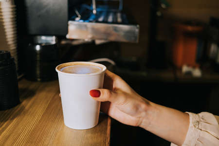 A paper coffee cup is on the counter in a coffee shop. The cup is taken by a woman's hand with red nails. Morning concept, coffee to go.の写真素材