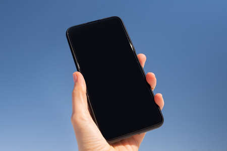 Smartphone with a black screen in a hand against a background of a bright blue sky. Copyspace.
