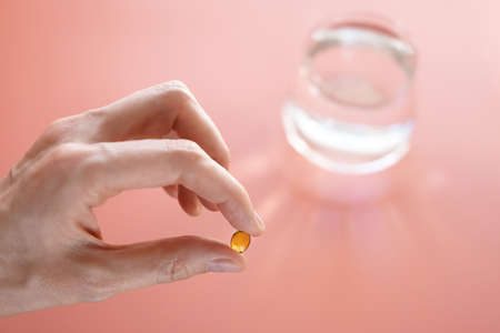 Vitamins of yellow color in a hand on a pink background. Behind - a glass of water. The concept of taking vitamins, dietary supplements, health promotion.の写真素材