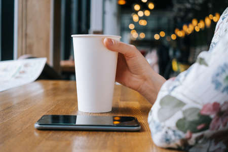 White large paper cup of tea on a table in a cafe. A woman's hand holds the cup, a smartphone lies next to it. The concept of a break, rest in a cafe. Copyspace.の写真素材