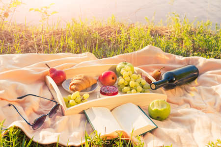 Picnic by the river. On a light-colored bedspread, a wooden tray with fruit, cakes and a bottle of wine. The light of the setting sun, a pleasant calm atmosphere, bright colors.の写真素材