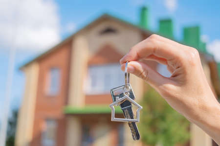 Close-up of a key with a house-shaped keychain in hand. In the background is a house. The concept of...