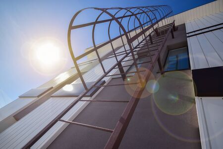 Beautiful brown metal staircase to the roof of a house in the sunlight against a white and brown wall and blue skyの写真素材