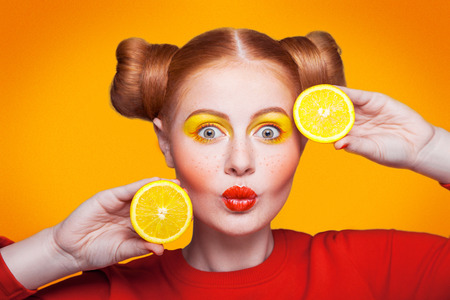 Young beautiful funny fashion model with lemon slice on orange background. with makeup and hairstyle and freckles.  studio shot, looking at camera with kiss.の写真素材