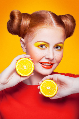 Young beautiful funny fashion model with lemon slice on orange background. with makeup and hairstyle and freckles. studio shot, looking at camera with smile.の写真素材