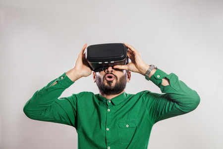 Young emotional middle eastern excited man with beard and green shirt using a VR headset and...