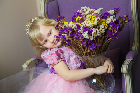 Little happy princess girl in pink dress and crown in her royal room sitting on chair and  holding vase with flowers.の写真素材