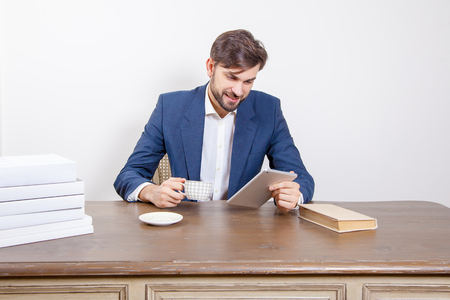Technology, people and business concept - handsome man with beard and brown hair and blue suit and tablet pc computer and some books and cup drinking coffee or tea sitting in the office holding tablet and smile. Isolated on white background.の写真素材