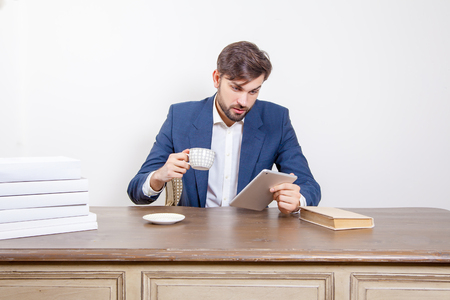 Technology, people and business concept - handsome man with beard and brown hair and blue suit and tablet pc computer and some books and cup drinking coffee or tea sitting in the office. Isolated on white background.の写真素材