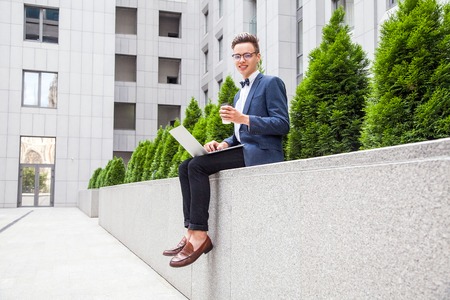 Attractive young businessman with laptop and coffee in hands on office building background. holding laptop and coffee and looking at camera.の写真素材