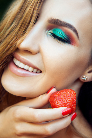 Young beautiful happy funny girl with red dress and makeup holding strawberry in summertime in the park. healthy lifestyle, diet beauty and happiness concept.の写真素材