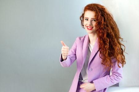 Portrait of successful happy beautiful business woman with red - brown hair and makeup in pink suit with thumbs up. looking at camera with toothy smile, studio shot on gray background.の写真素材