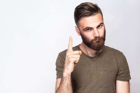 Portrait of serious bearded man with warning finger and dark green t shirt against light gray background. studio shot.の写真素材