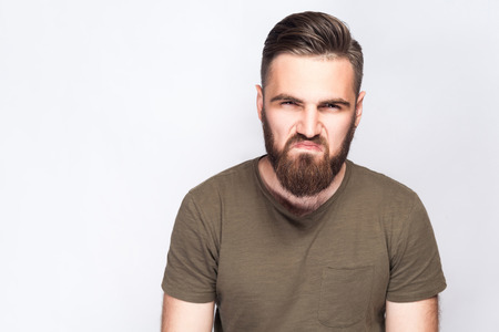 Portrait of angry sad bearded man with dark green t shirt against light gray background. studio...
