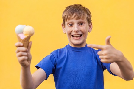 Blonde beautiful boy with freckles and blue T-shirt showing  ice cream with fingers and looking at camera with surprised face. Studio shot, isolated on a yellow background.の写真素材