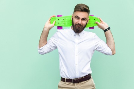 Bearded young hipster looking at camera, holding penny skateboard under his head. Light green background. Studio shotの写真素材