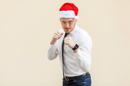 Boxing. Young adult ginger businessman on red santa hat, ready for fight on light orange background. Studio shotの写真素材