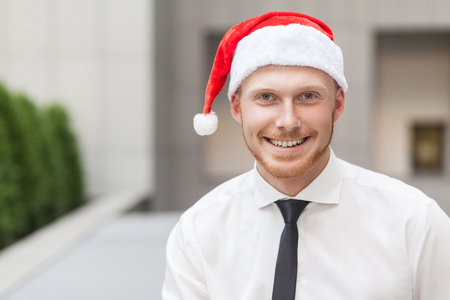 Portrait of success ginger businessman on santa hat. Looking at camera and toothy smile. Focus on face. Outdoor shotの写真素材