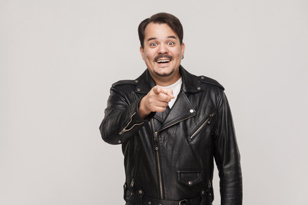 The happiness man, wearing  black jacket, having happiness looks, pointing finger at camera and toothy smiling. Isolated studio shot on gray backgroundの写真素材