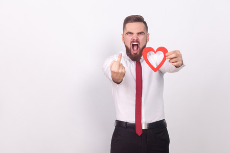 Fuck you, love! Angry man showing hate sign, holding heart. Indoor, studio shot, isolated on gray backgroundの写真素材